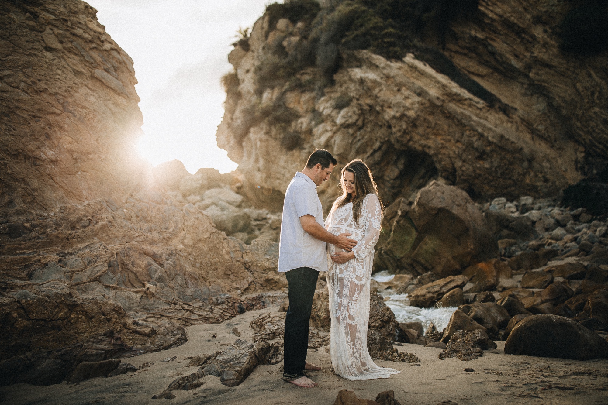 young pregnant couple on rocky beach with sunset behind them